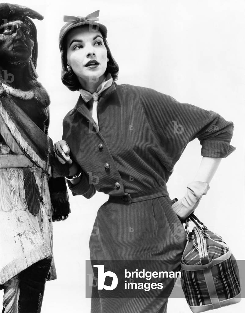 Corduroy shirtdress with dolman sleeves, a narrow skirt, and self belt. The model stands next to a wooden Indian and carries a plaid fabric purse with leather trim and a zipper closing. Fall 1951