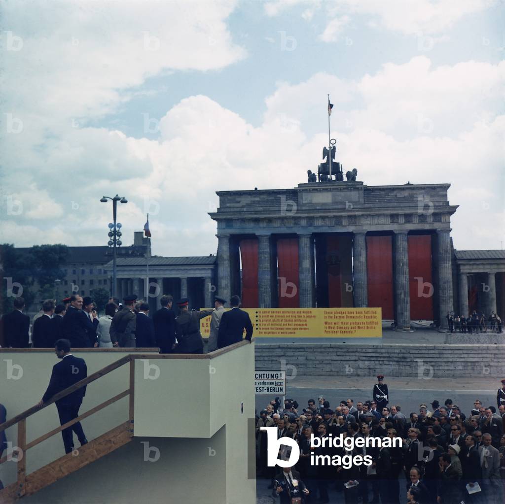 President John Kennedy, on a platform overlooking Berlin Wall and Brandenburg Gate in Berlin. The East German government decorated the gate with a flag and constructed a yellow sign justifying the wall to prevent German aggression. June 26, 1963