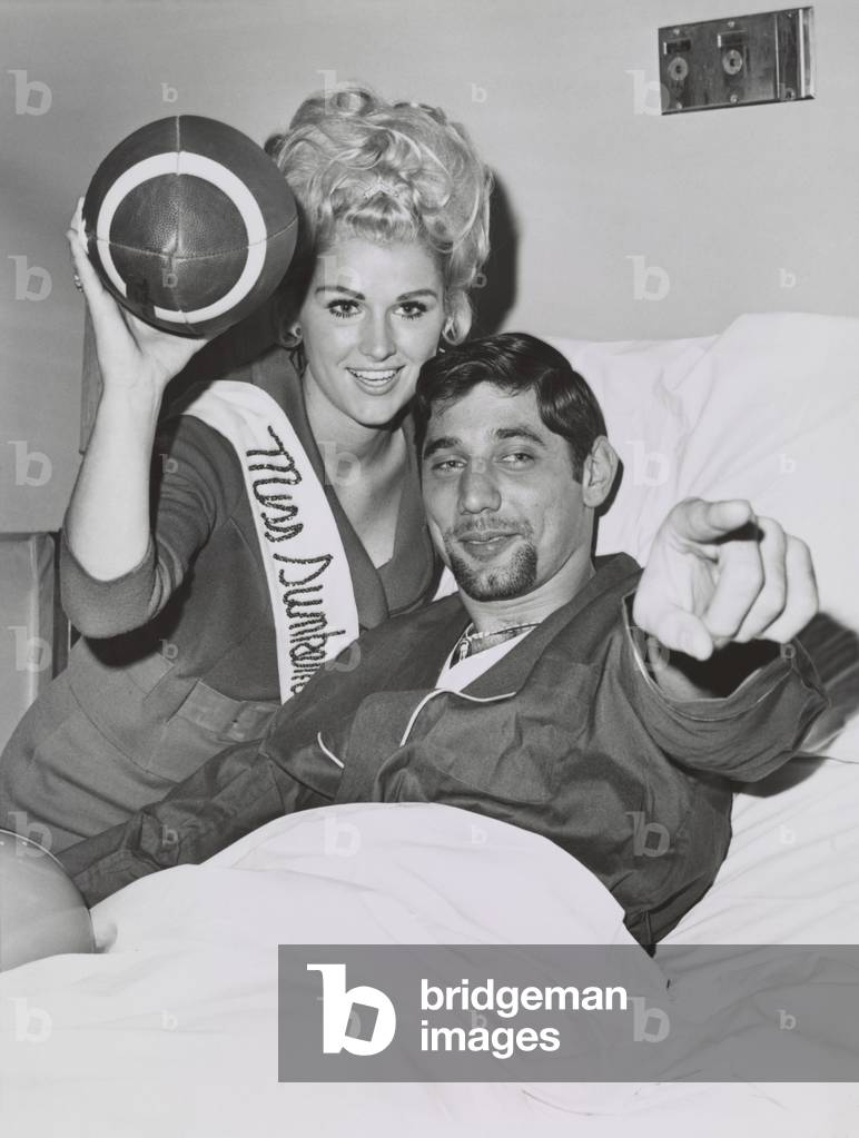 Joe Namath, New York Jet's quarterback, in a hospital bed awaiting an operation on his right knee. The blond keeping him company is Miss Sunken Gardens, of Petersburg, Florida. Dec. 28, 1966.