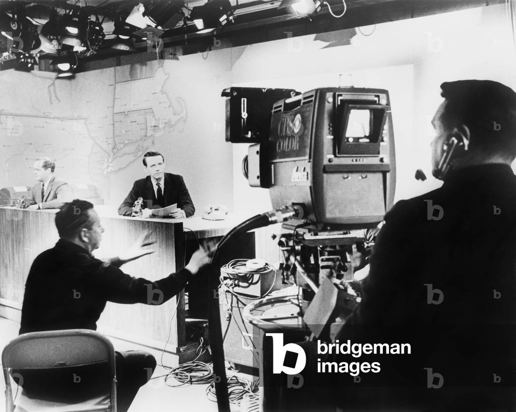 Under hot studio lights, a CBS News anchor performs under the director of his producer (seated between anchor and camera), for a color television camera in 1967