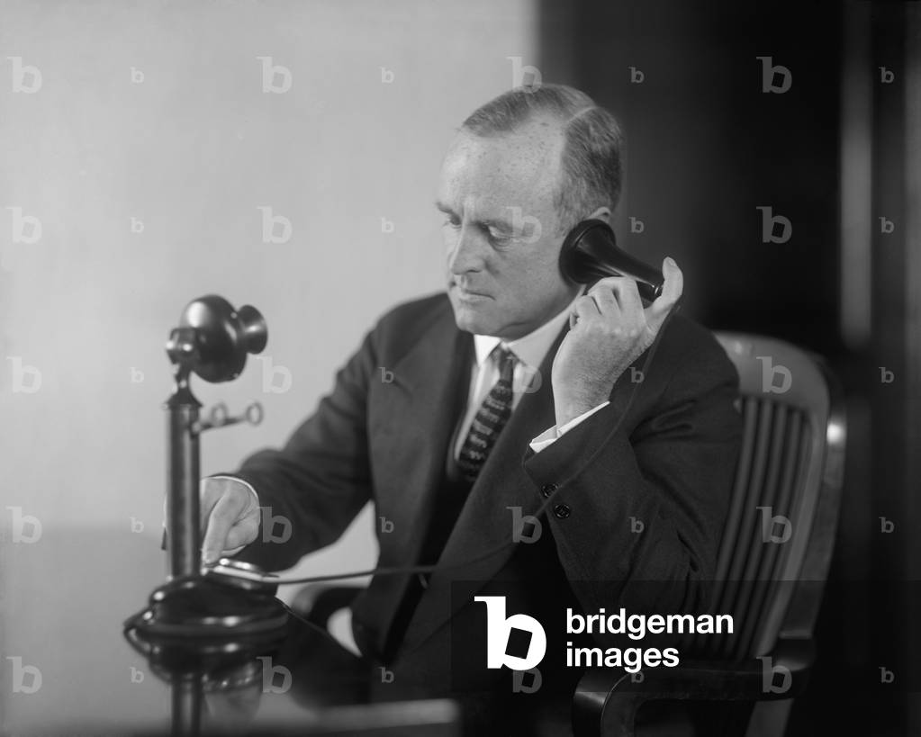 Businessman dialing an early 20th century desk phone with separate ear receiver and mouth transmitter. c. 1920