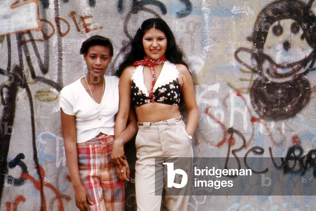 1970s Americ. Two young girls pose in front of a graffitied wall in Brooklyn, New York City, c. 1973