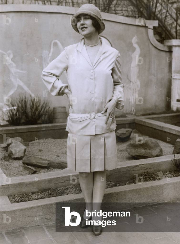 1927 suit with a mid-knee pleated skirt and matching waistless, hip-belted jacket. The outfit is toped by a cloche hat. March 1927