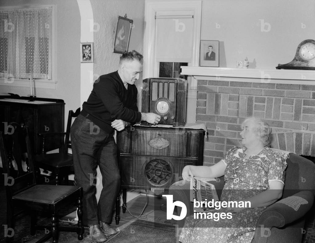 A husband tunes the radio while his wife holds populist right-wing radio priest, Father Coughlin's newspaper SOCIAL JUSTICE, with a headline, STALIN ORDERS WORLD REVOLUTION. This comfortable middle class home was in Royal Oak, Michigan, where Father Coughlin was a priest. 1938