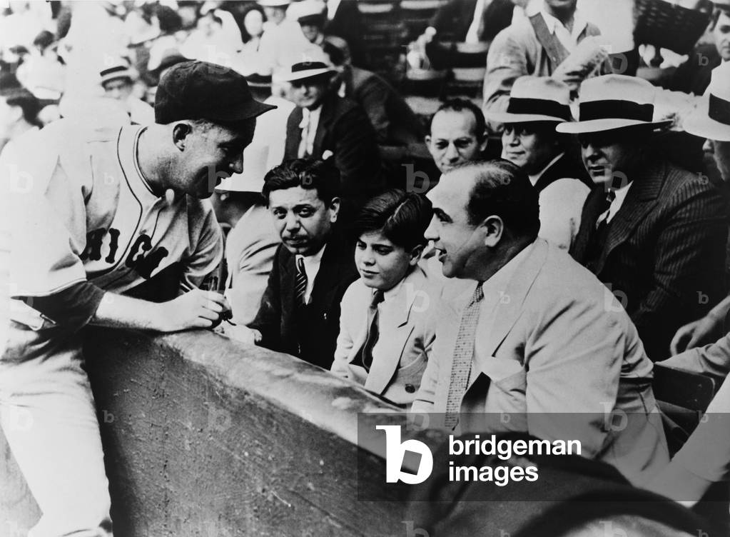 Chicago Cubs player Gabby Hartnett autographing a baseball for Sonny Capone who is sitting with his father Al Capone and other gangsters at a 1931 charity baseball game