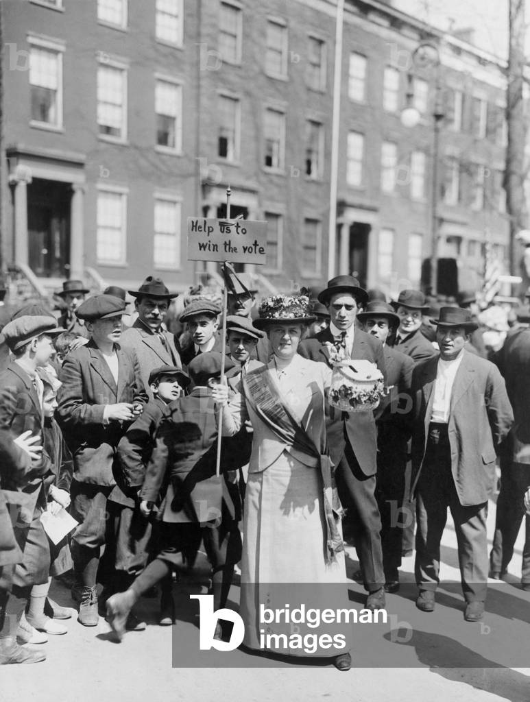 Women identified as Mrs. Suffern, is surrounded by a crowd of men and boys, while she holds a home-made banner in women suffragist parade 'Help us to win the vote.' 1914