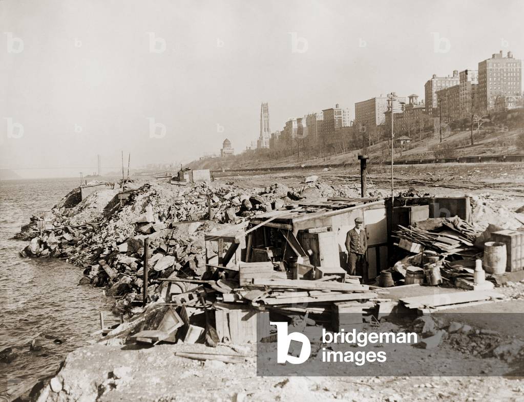 An unemployed a man standing near his hut in a New York City shanty town during the Great Depression. In the background are Upper West Side landmarks, Grant's Tomb and Riverside Church. 1933