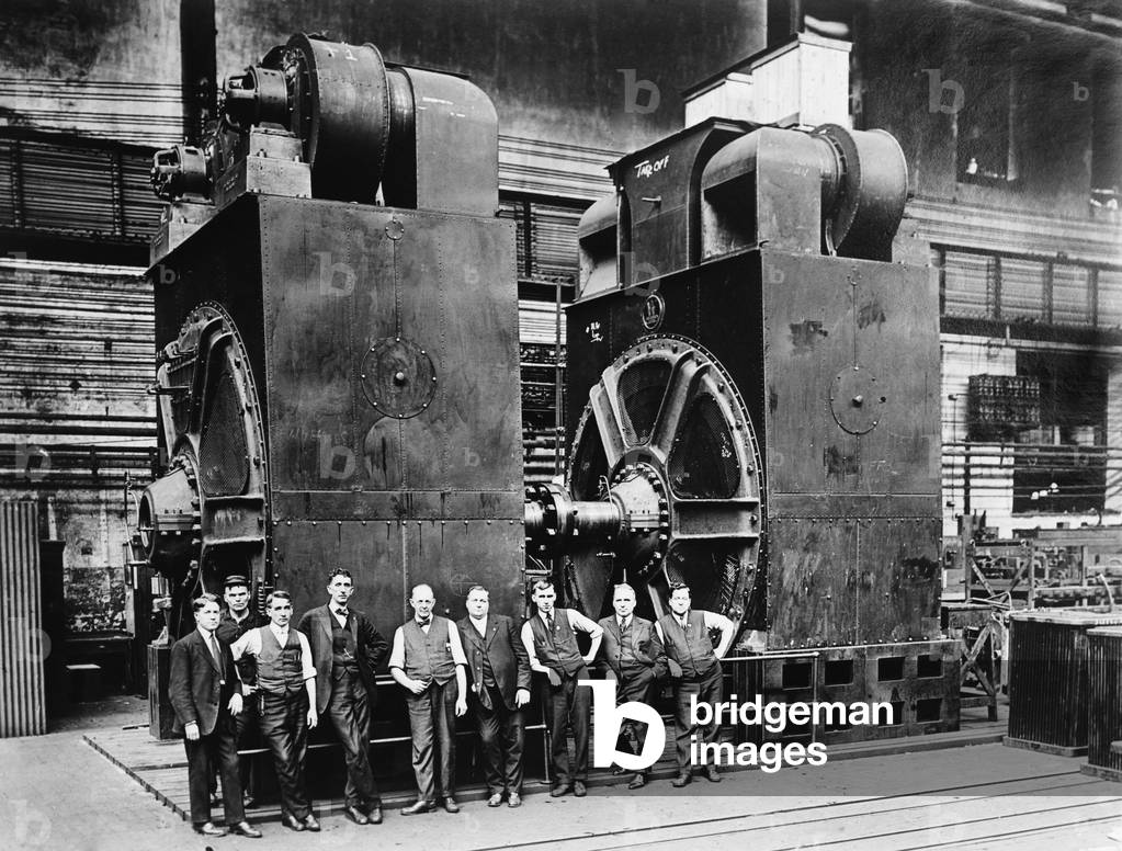 Nine men in suits are dwarfed by two massive dynamos, (electric generators) in a factory. c. 1920