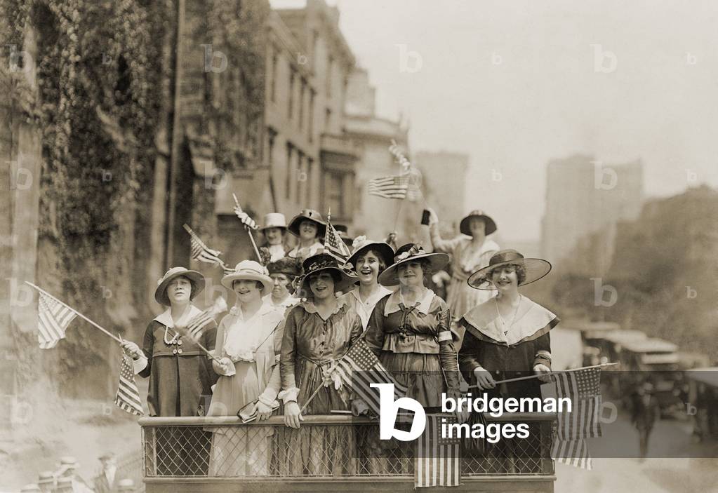 Smiling young women waving flags from the top of an open vehicle in a New York City parade. c. 1910-1915