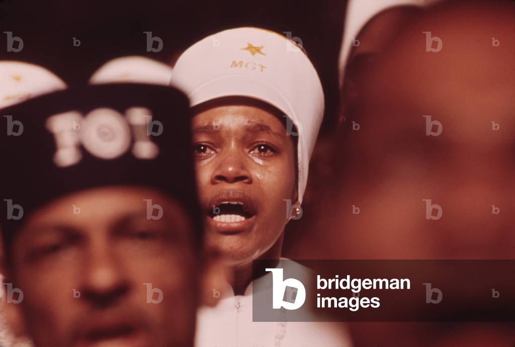 A Black Muslim woman reacts to Elijah Muhammad's Annual Savior's Day message. The FOI on the cap of the man in front indicates he is a member Fruit of Islam a paramilitary group. March 1974