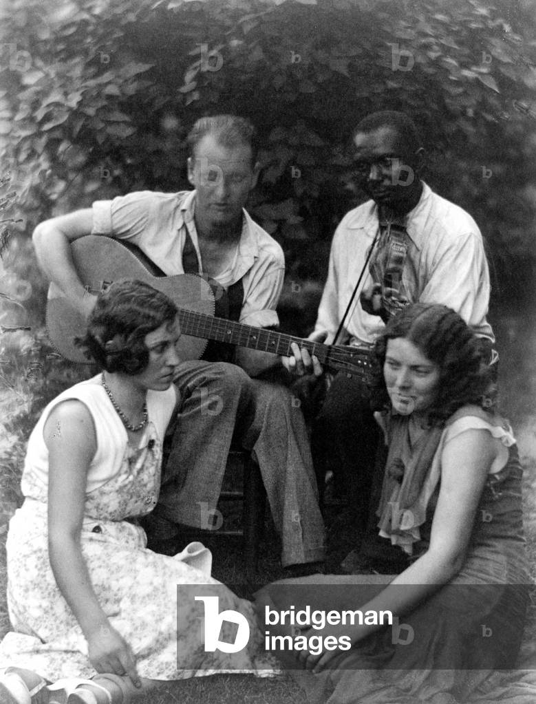 Folk Music. Four musicians including a man playing a guitar, a man playing a violin, and two women seated on ground, c. 1930