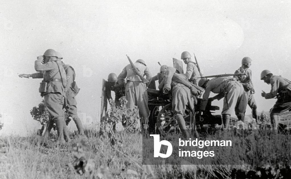 A squadron of Italian soldiers, with rifles on their backs, are preparing and aiming a cannon to be fired against the enemy under the directions of captain Tognetti who, together with other soldiers, is observing the battle field. It is the afternoon of July 14, 1938, during the action known as 'Del Levante' (b/w photo)