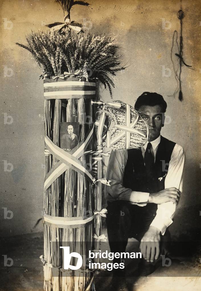Antonio Floridi posing near fasces of lictors made of ears of wheat. Farm owned by Camillo Frizzoni, in Morengo, province of Bergamo. On top of the fasces is a photo of Mussolini (b/w photo)