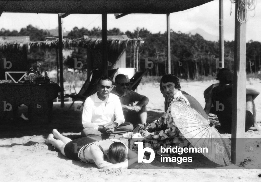 Portrait of a group of people seated on the beach, under a parasol shed roof. In the background a wood can be glimpsed, Versilia
