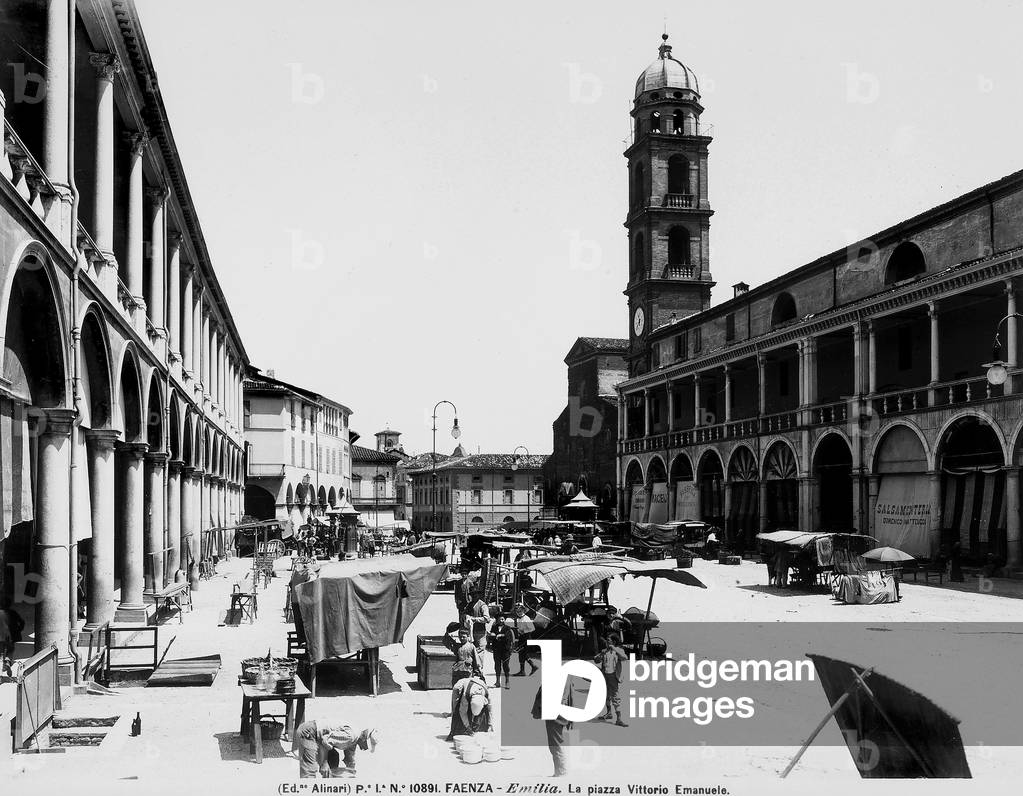 View of the busy Piazza Vittorio Emanuele (today Piazza del Popolo e Piazza della Libertà) in Faenza, occupied by some temporary commercial stalls. On the right and on the left, two buildings with galleries and arches on each level.