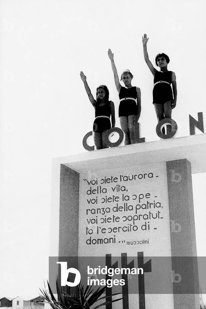 Three little girls do the roman greeting standing up on the entrance arc of a summer colony in Marina di Pietrasanta. On the arc Mussolini's words inviting kids to become the future soldiers of the country (b/w photo)