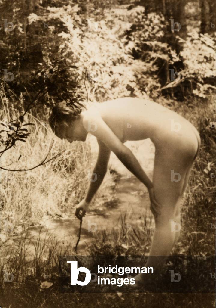 A young woman, nude in the woods, seen in profile as she bends over to put a branch in a stream.