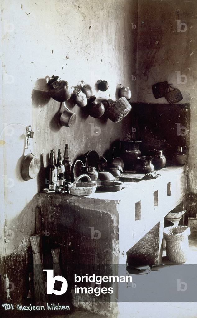 Corner of traditionally furnished kitchen in a Mexican home: the kitchen as such is composed of a brick structure with openings at the front, into which the hot coals were inserted, to be spread out on the flat top horizontal plane, where the food was heated and cooked. On such a structure, which occupies the whole of the corner in the photograph, much crockery can be seen, some hung on the walls