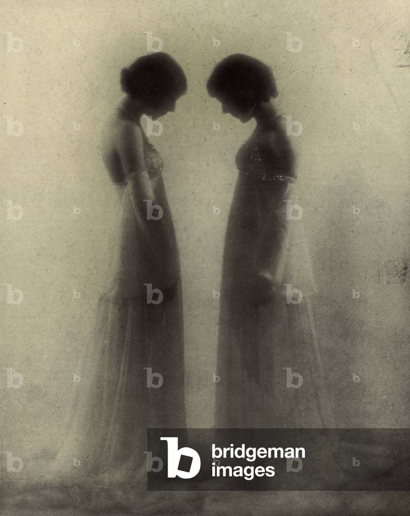 Full-length portrait, in profile, of two girls. The lovely young women are dressed in identical elegant white evening dresses and have their hair gathered up