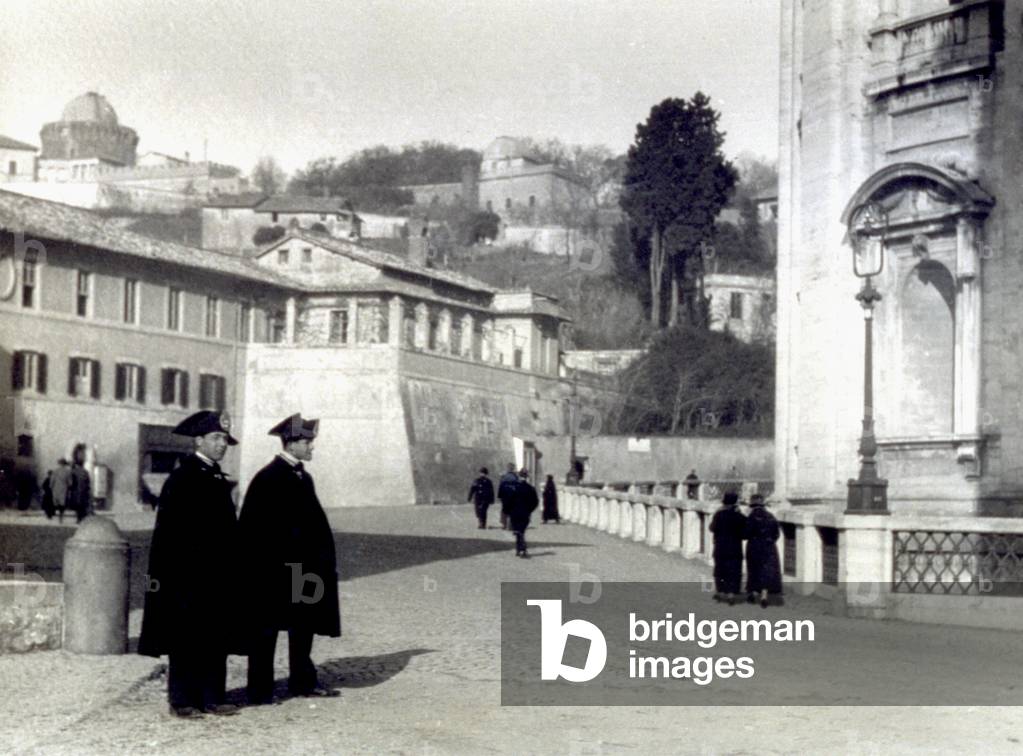 A street in Rome, with people out walking. In the foreground two Carabinieri in uniform