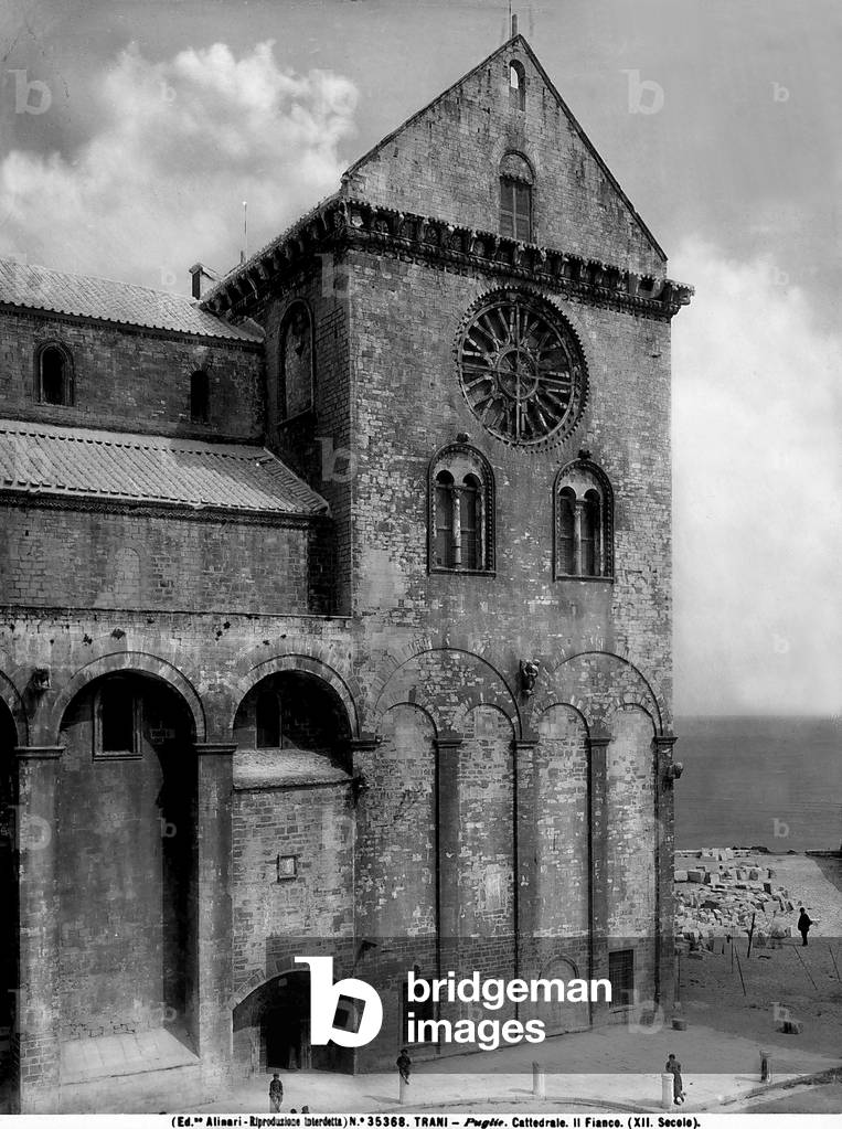 View of the right side of the Cathedral of Trani. The high transept is adorned with a rich cornice with figured brackets, in the lower part, two pairs of blind arches, two windows with two lights and a large rose window are visible. Some people are walking nearby.
