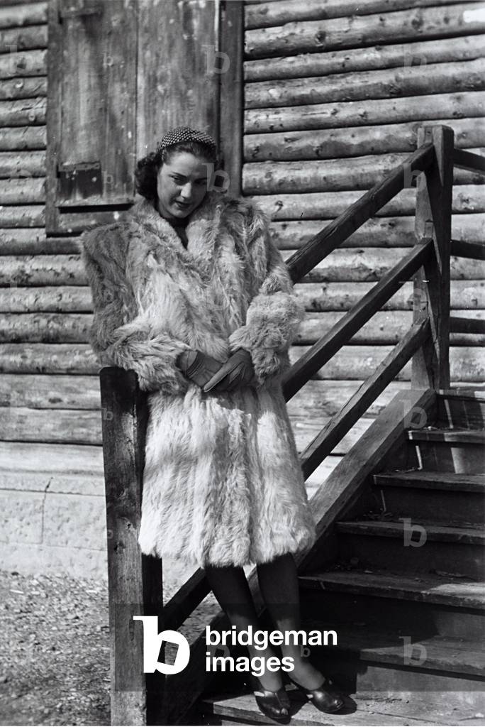 A model posing on the steps of a mountain chalet wearing a light colored fur. (b/w photo)