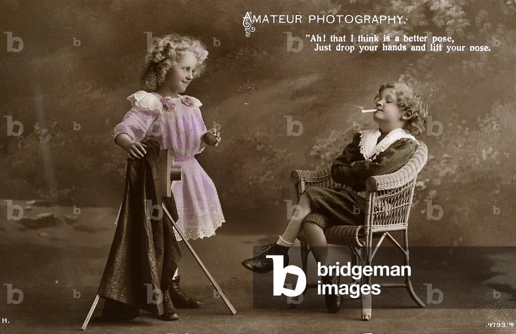 A boy sitting in a chair, arms folded and cigarette in mouth, takes directions on how to pose from a very young female photographer