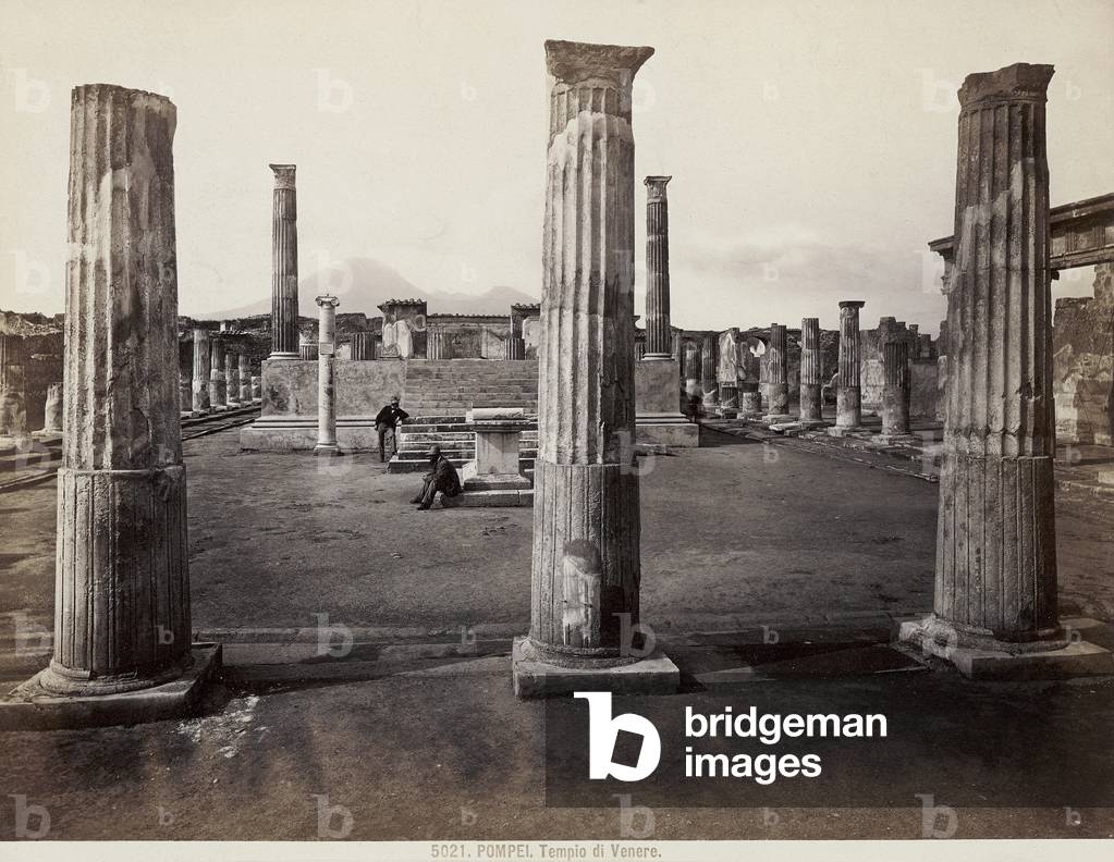 The 'Temple of Venus' in Pompeii. In the foreground the remains of columns; at the center of the sacred area stands a high podium preceded by a flight of steps. Two men can also be seen in the shot
