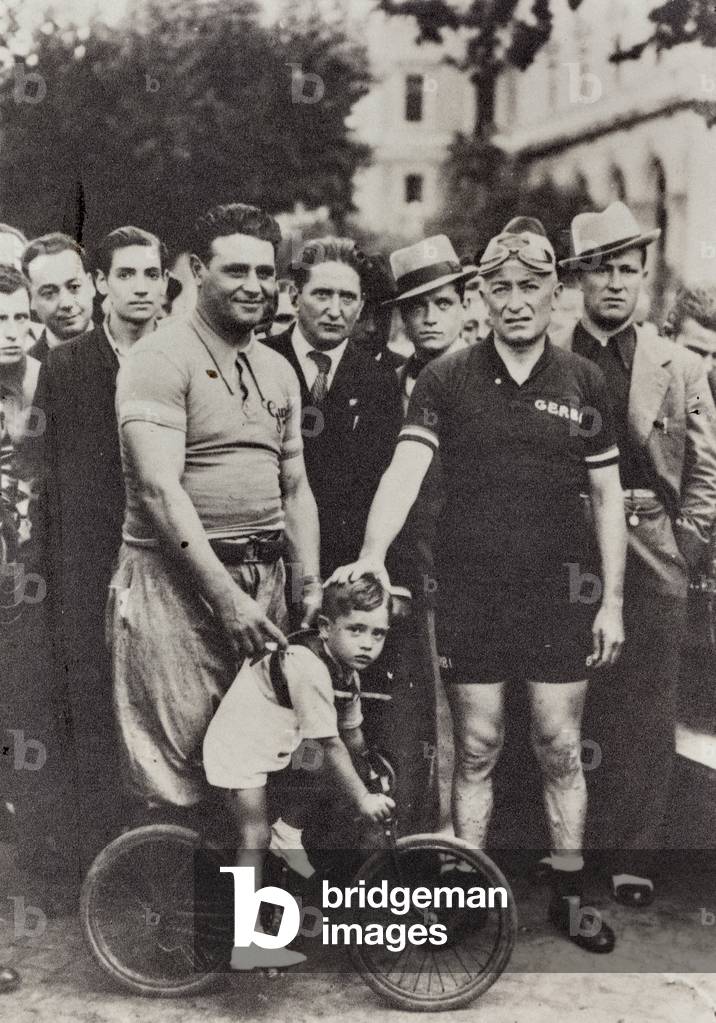 Two cyclist pose with a little boy riding a small bike; behind them, a group of men.