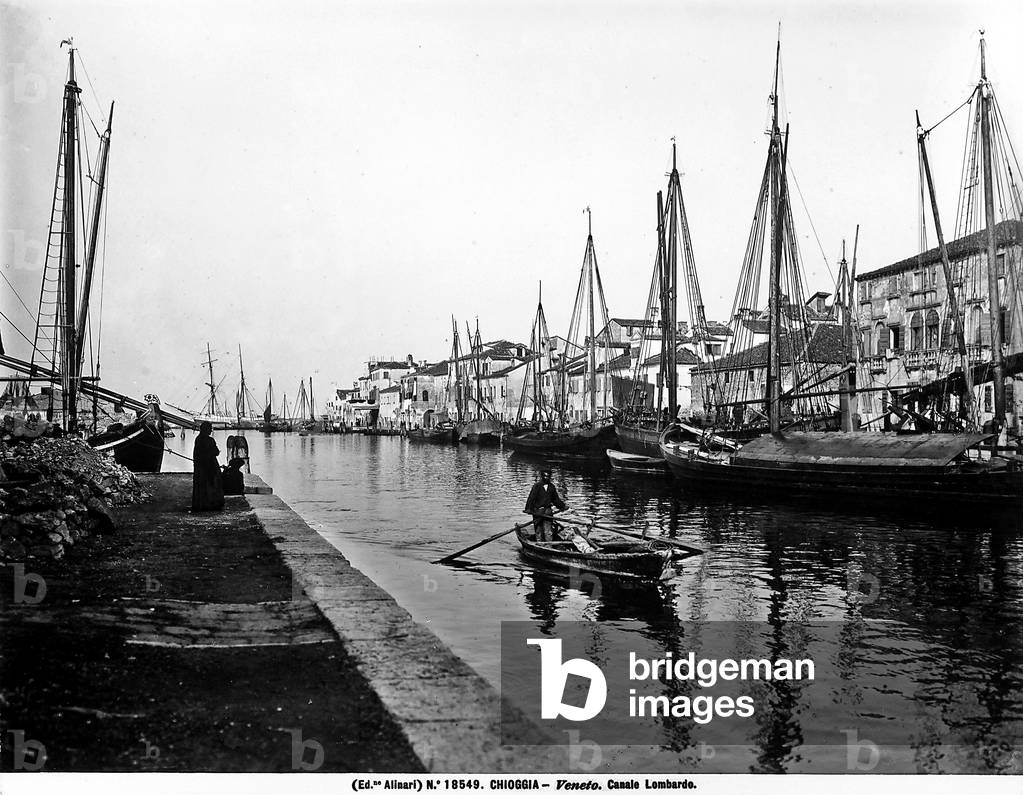 View of the Lombardo Canal in Chioggia. Many sailboats can be seen along the canal; in the center, there is a man rowing a small rowboat