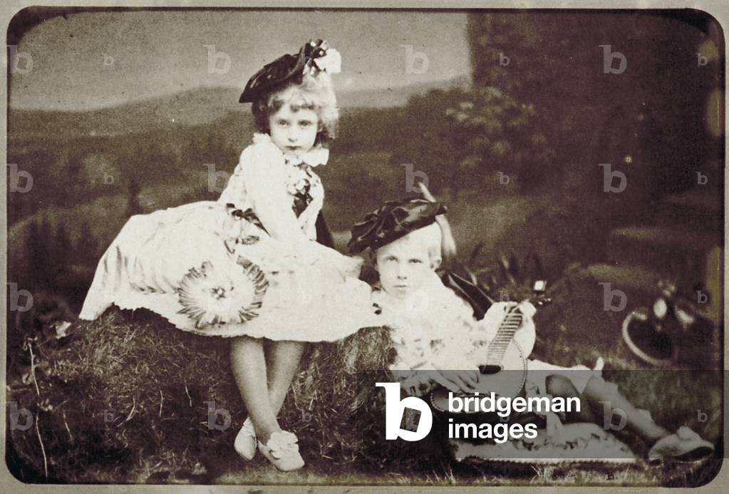 Victor Emmanuel III and princess Trivulzio as children, in costumes. The scene is set in a country ambience. The two children are seated on the grass. Victor Emmanuel III holds a mandolin