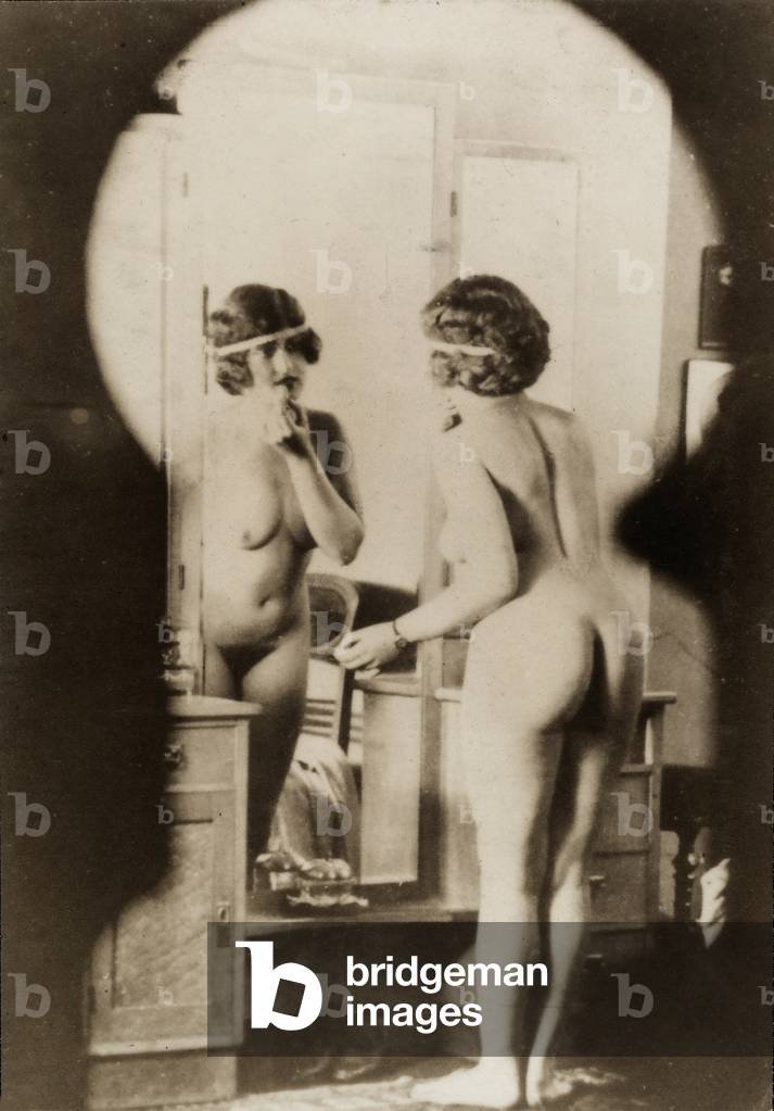 Nude full-length portrait of a young woman in front of a mirror, seen from a keyhole.