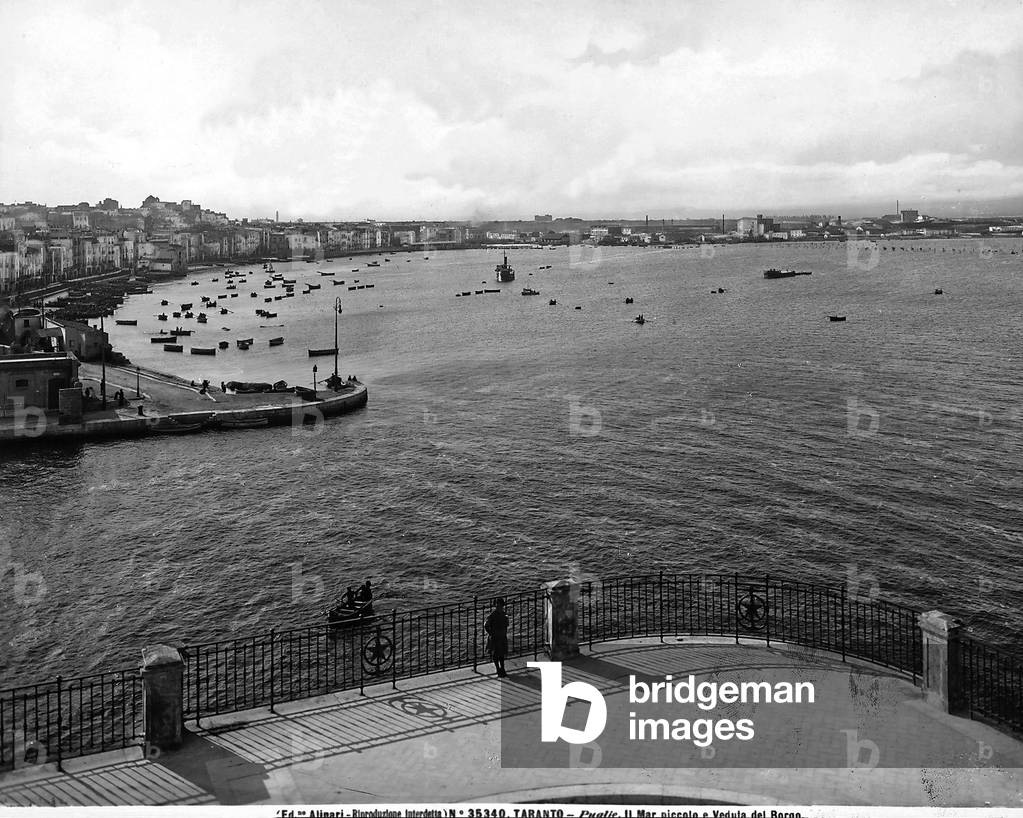Panoramic view of porto di Taranto; in the background some industrial establishments are visible, on the left are moored boats and a view of the city, in the foreground, a woman is leaning on the railing of piazzale, observing the view