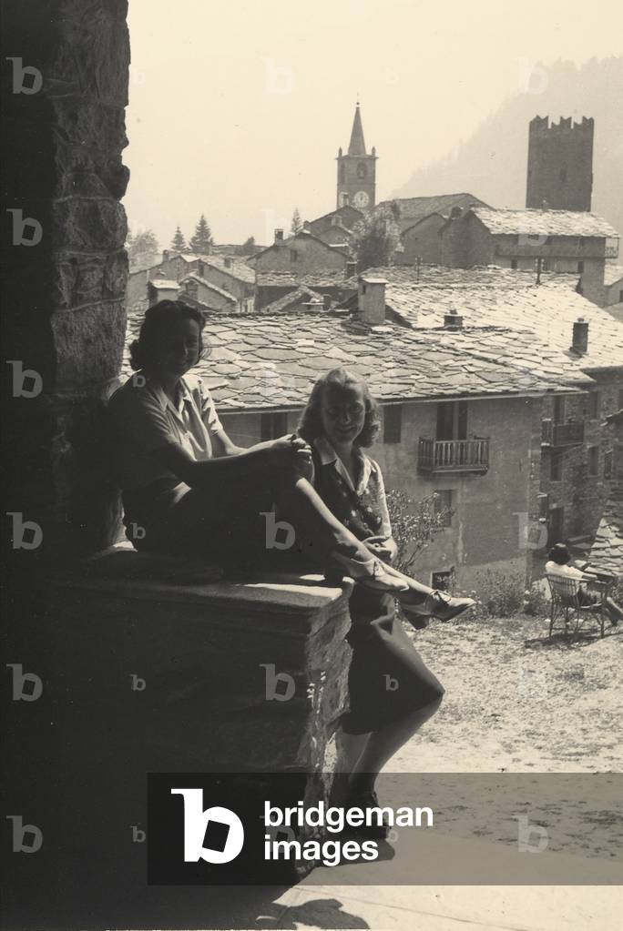 Two young girls rest on a stone wall. In the background, a mountain village, 1940-50 (b/w photo)