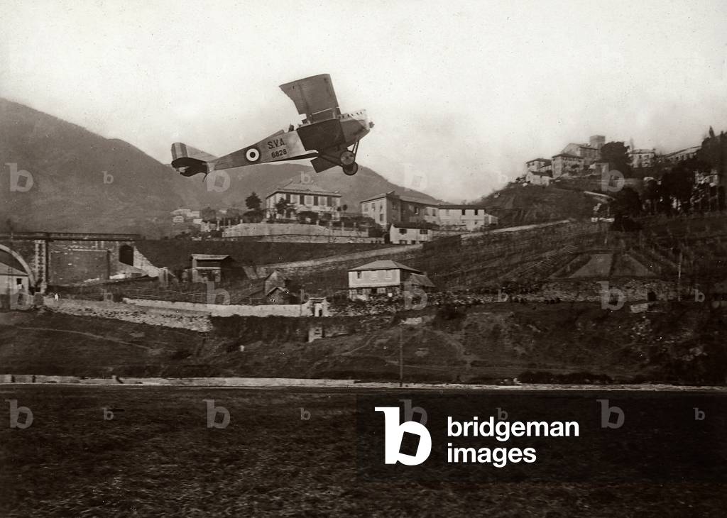 An S.V.A. warplane flies over a mountain village, in the background a bridge is visible. (b/w photo)