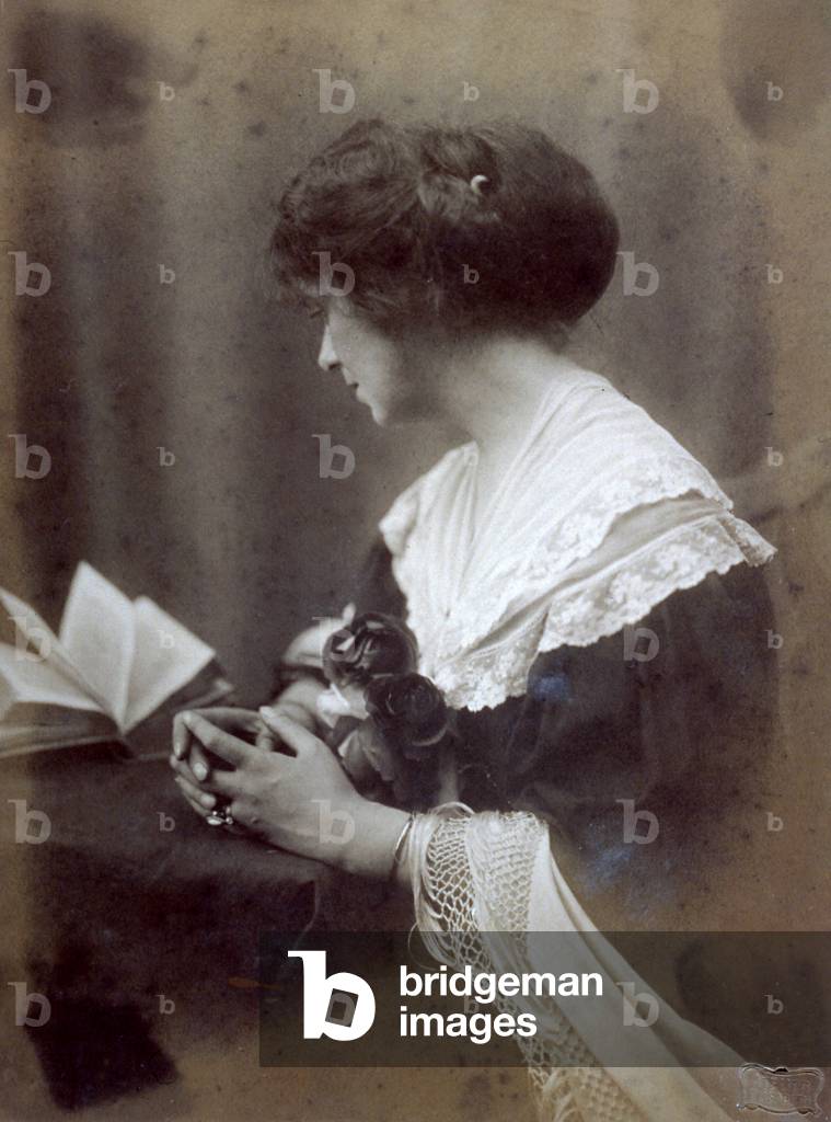 Three-quarter-length portrait of an elegant young lady sitting at a table. She is sitting in profile, with her face turned to a book in front of her