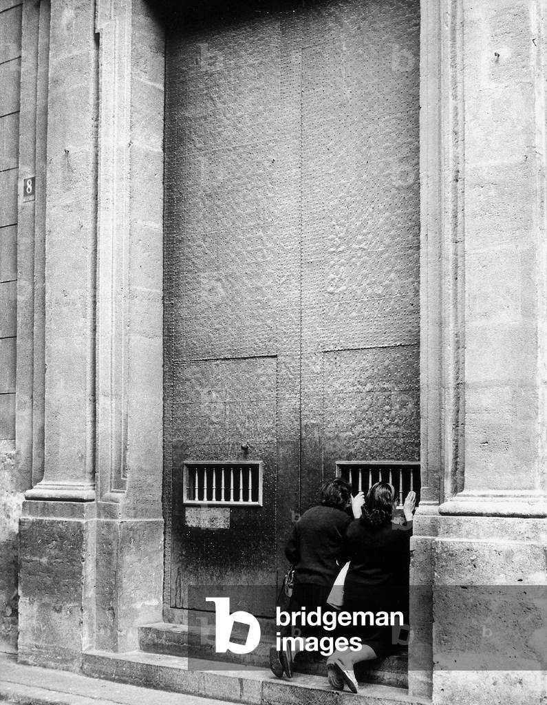 Two women peek through the bars of the main doors, at the inside of a building.