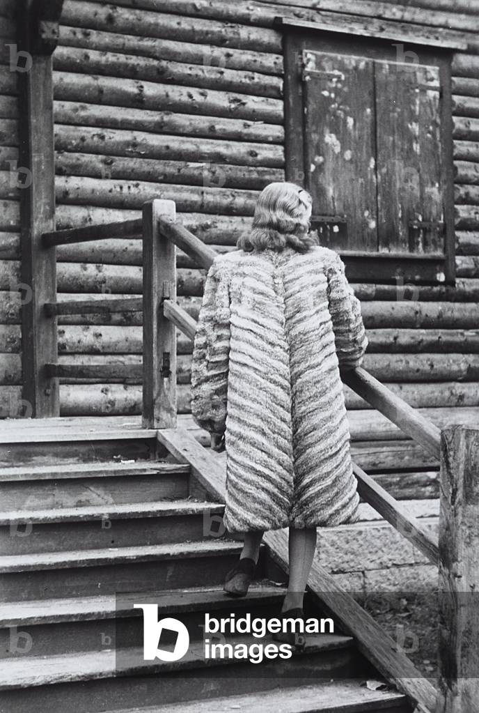A model posing on the steps of a mountain chalet wearing a light colored fur with dark stripes with her back to the camera. The 