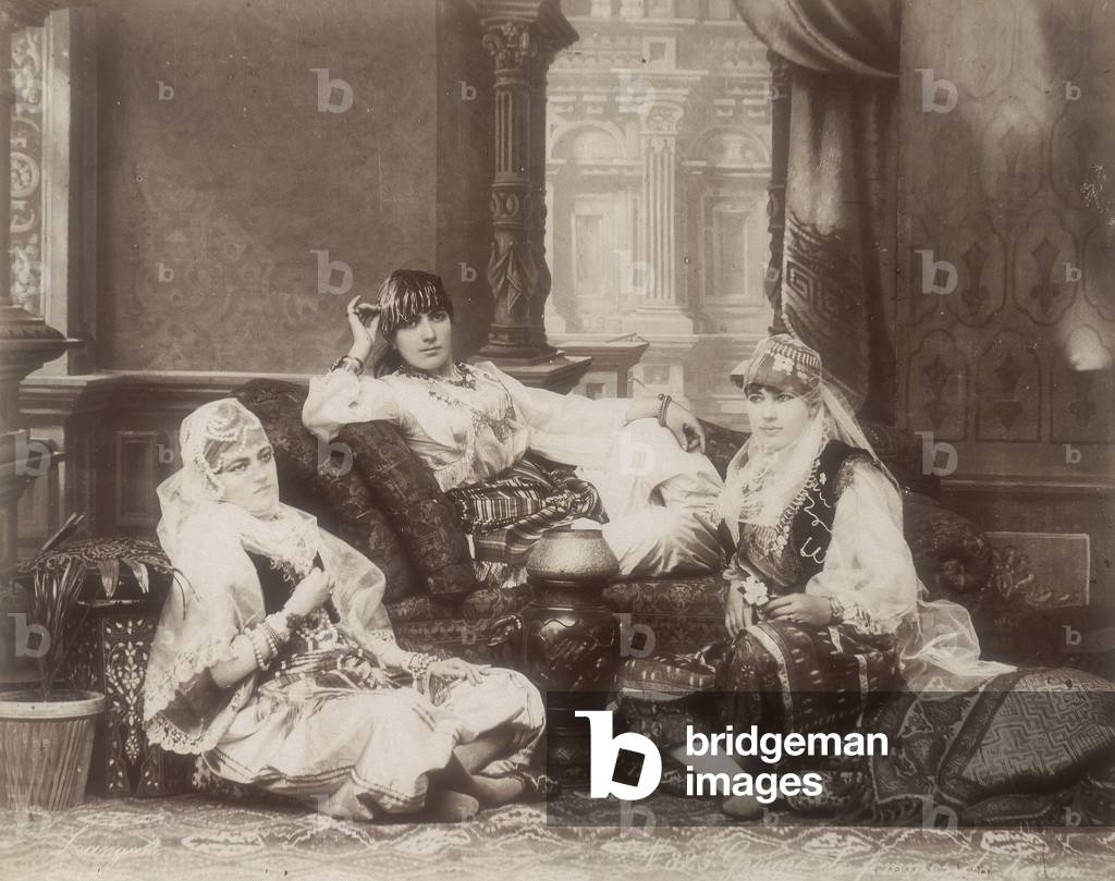 Three young egyptian women in ethnic dress. One of them is reclining on a sofa. At her feet, on soft cushions, the other two girls are posing, their faces covered by veils, 1870 (print on double-weight paper)