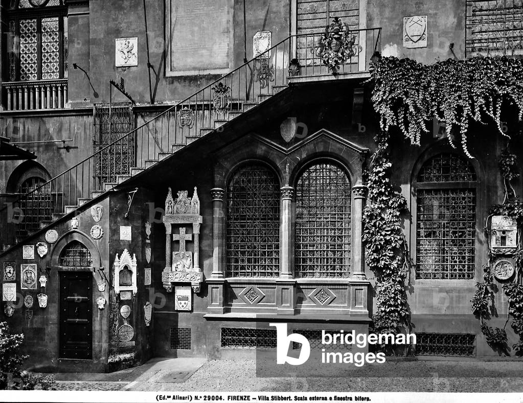 View of the external staircase of Villa Stibbert in Florence. Under it, two curved windows covered in iron and framed by columns and gables forming a mullioned window with two lights.