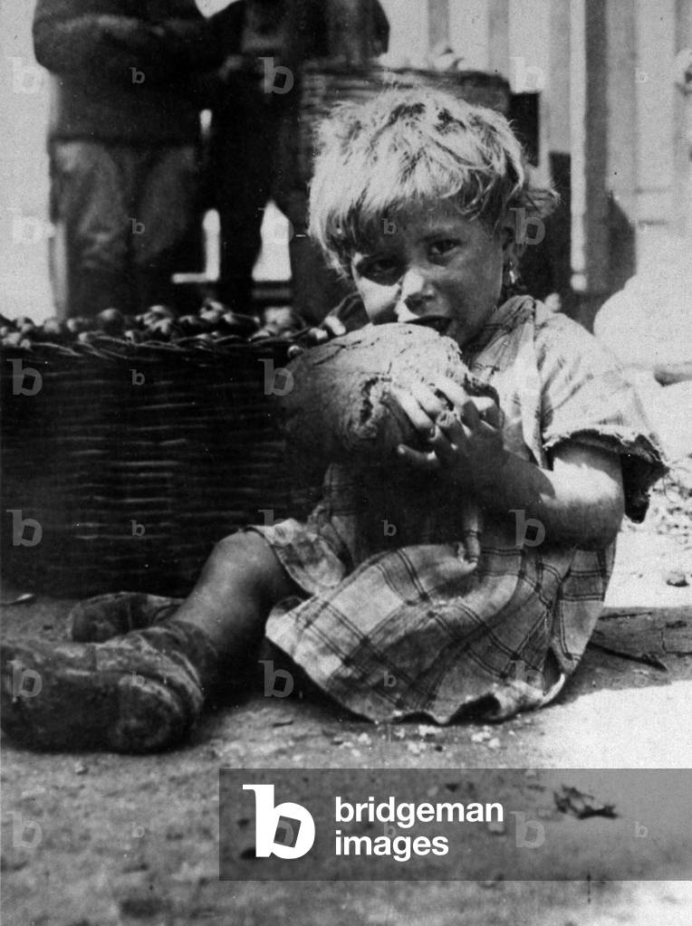 Portrait of a little girl. She is dirty and poorly dressed and is seated on the ground eating a large piece of bread