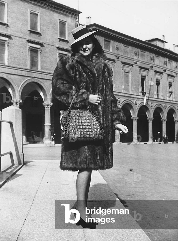 A model posing in a square with porticoes wearing a creation by the furrier Sarti. She is wearing a wide-brimmed hat and a purse made of crocodile skin. (b/w photo)