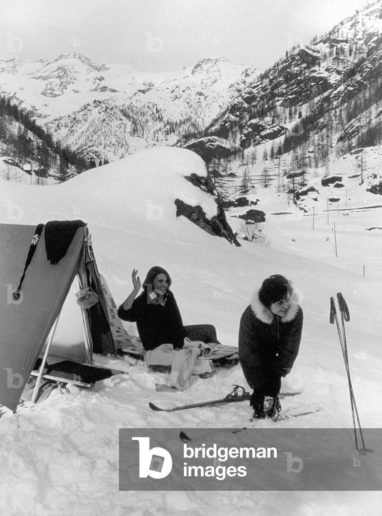 Portrait of two women next to a tent in a mountain landscape, covered with snow. One of the two women is shown putting her skis on, the other is sitting on a small bed
