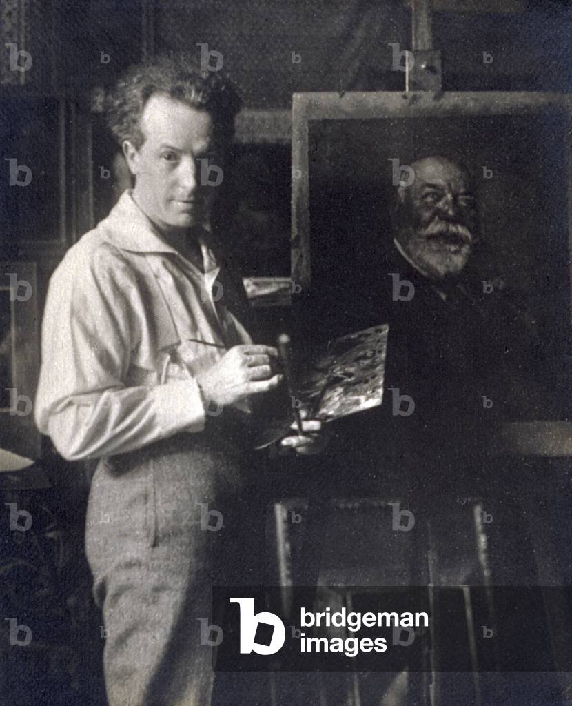 Three-quarter length self-portrait of the photographer Cesare Scarabello. He is posing before a painting of the face of an old man, set on an easel, holding the palette and a brush and looking towards the camera (b/w photo)