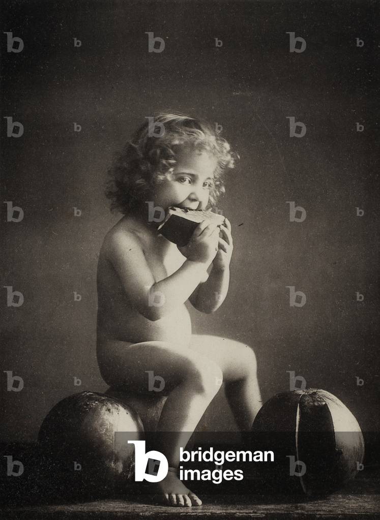 Full-length portrait of a small child, nude and seated on a watermelon, eating a slice of melon