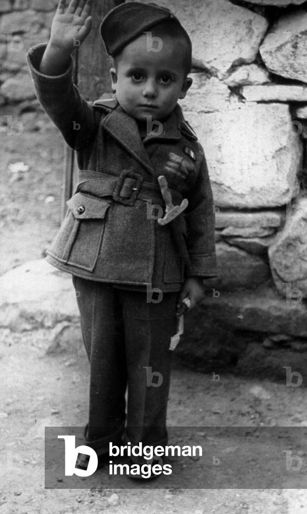 Full-length portrait of a child dressed as a soldier, complete with a small wooden sword, he is gravely making a Fascist salute (b/w photo)