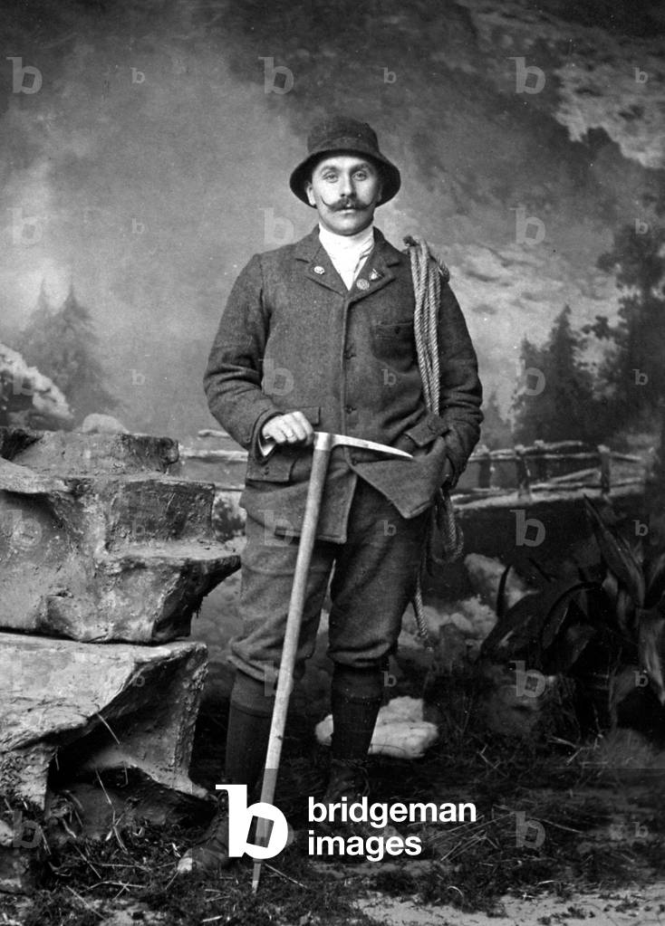 Full-length portrait of a man dressed as a mountain climber. He is leaning on a pick and has a rolled up rope on his left shoulder. Behind him a mountain backdrop
