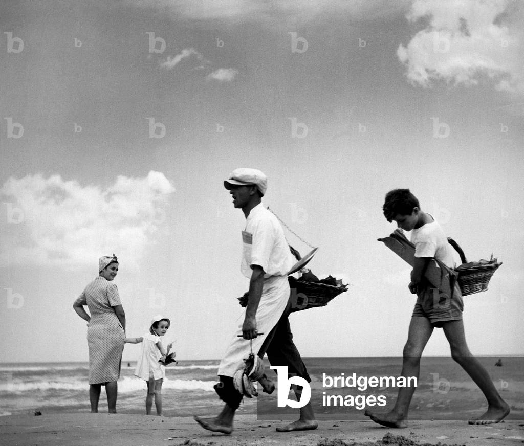 Two travelling salesmen walking on a beach. In the background there is a woman and a baby.