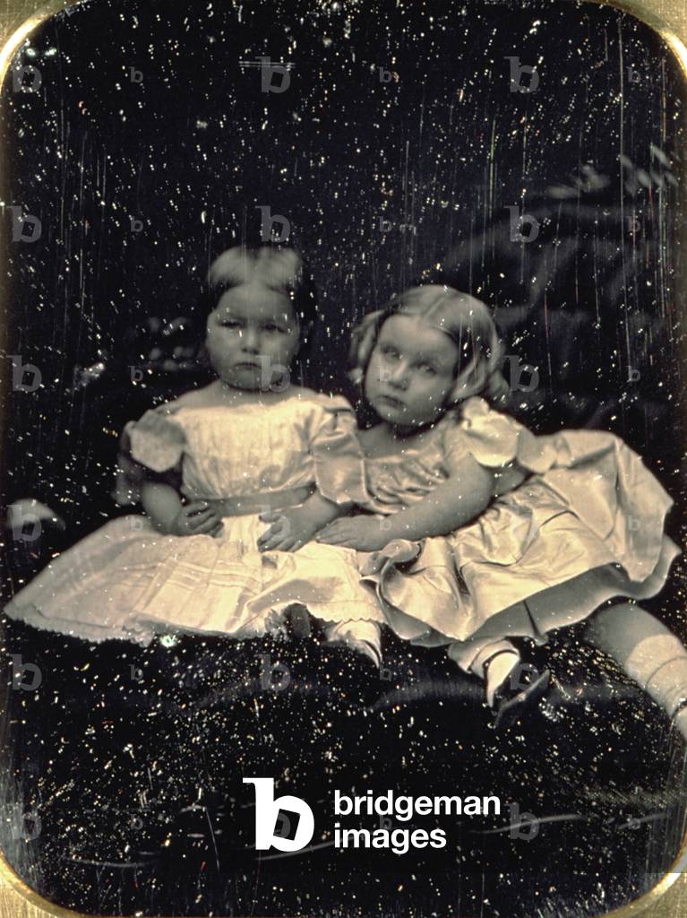 Portrait of two little girls in elegant summer dresses of romantic period. They are posing in the studio, seated on a sofa, and embracing
