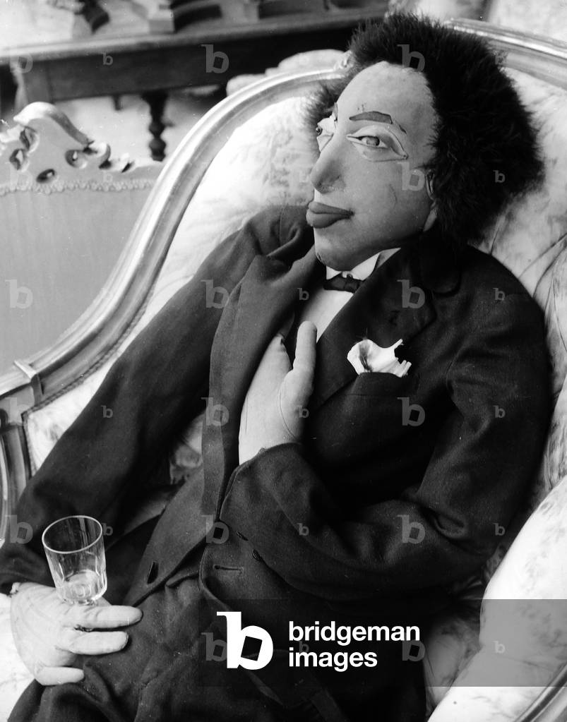 A dummy depicting an elegantly dressed man on a chair with a glass in his hand.